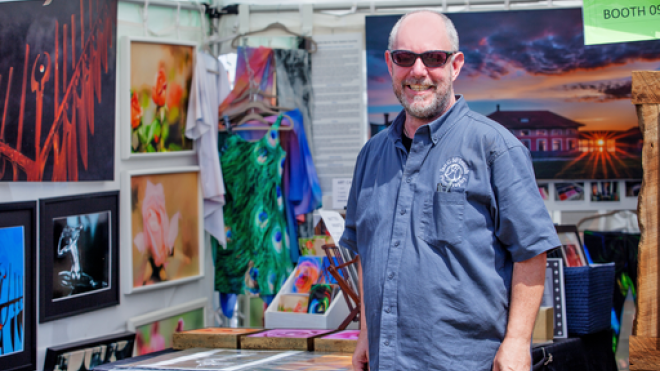 Man holding art supplies standing in room surrounded by paintings.