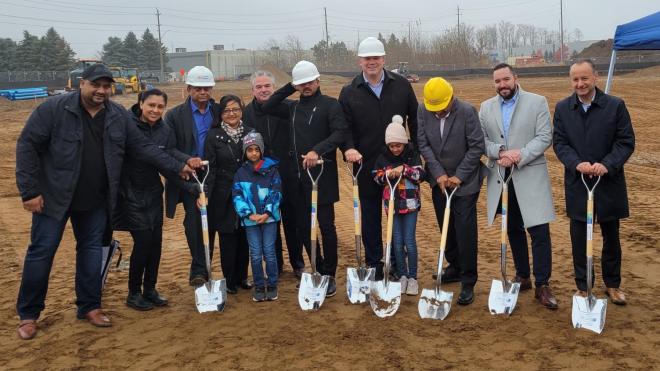 Row of people posing for a ceremonial groundbreaking. They are wearing white and yellow construction helmets and are holding silver spade shovels.