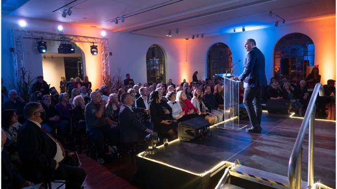 Person addressing seated audience in a large room. The subject is standing behind a podium with microphones on it.