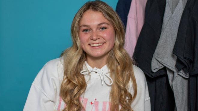 Young person wearing a custom hoodie with a clothing rack with t-shirts in the background
