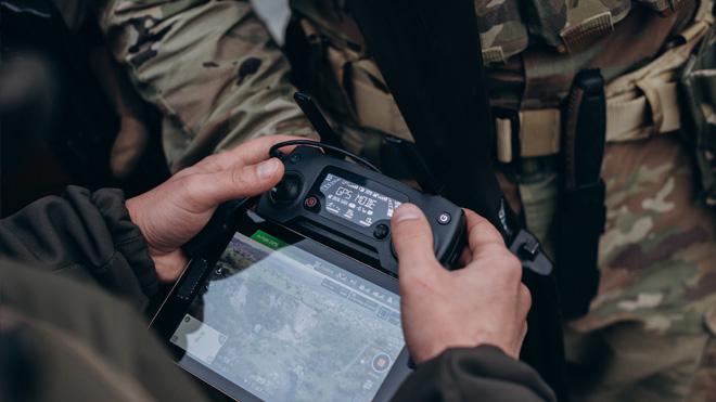 Hands hold a drone controller with an aerial map screen, soldiers in camouflage nearby.