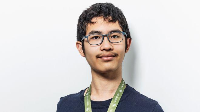 Young person with glasses and a lanyard, wearing a dark shirt, smiling slightly.