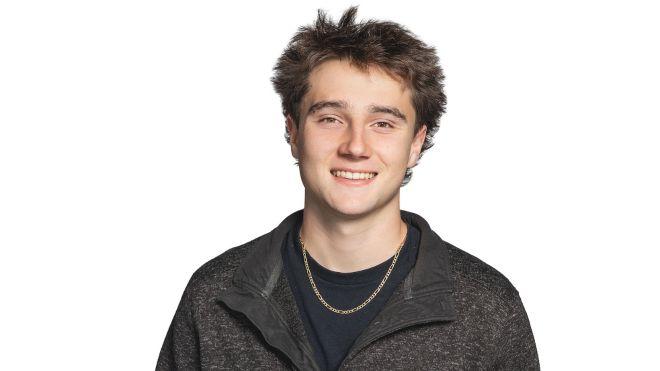 Young man smiling, wearing a black jacket and chain necklace, on white background.
