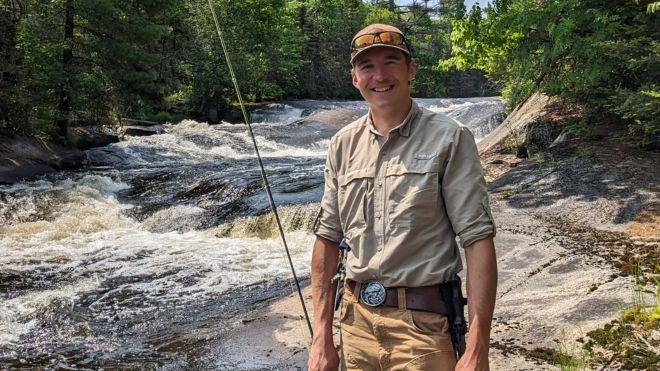 Man smiling by a river, holding a fishing rod, surrounded by trees.