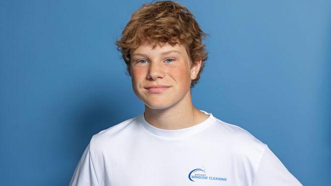 Teen boy in white shirt standing against a blue background, smiling softly.