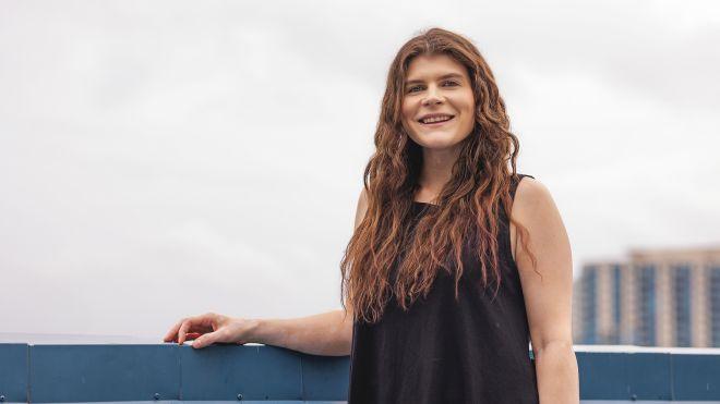 Smiling woman with long hair, wearing a black top, stands outdoors by a blue railing.