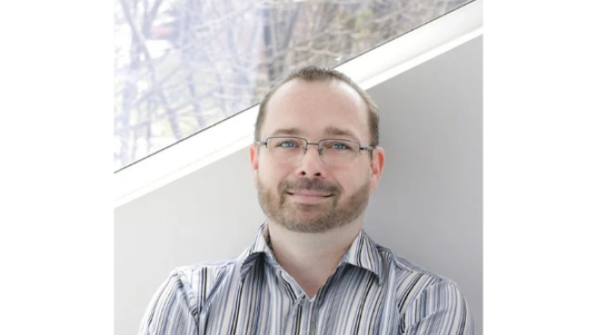 Man with glasses and a striped shirt, standing in front of a window.