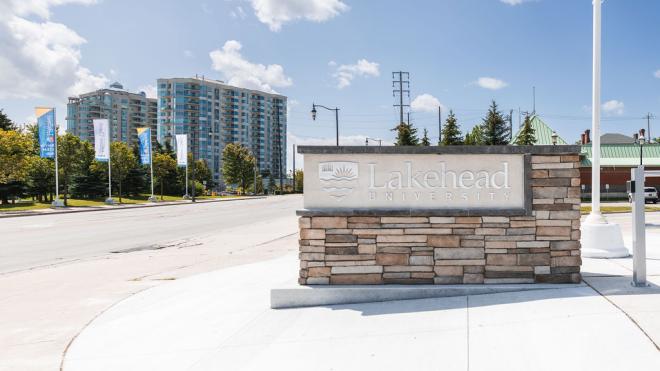 Stone Lakehead University signboard by a road, trees, and a tall building in the background.