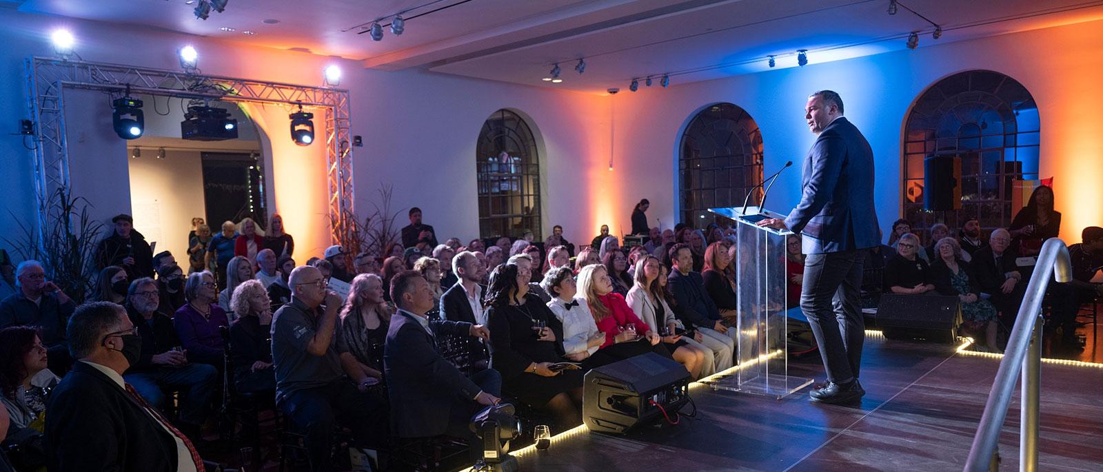 Person addressing seated audience in a large room. The subject is standing behind a podium with microphones on it.