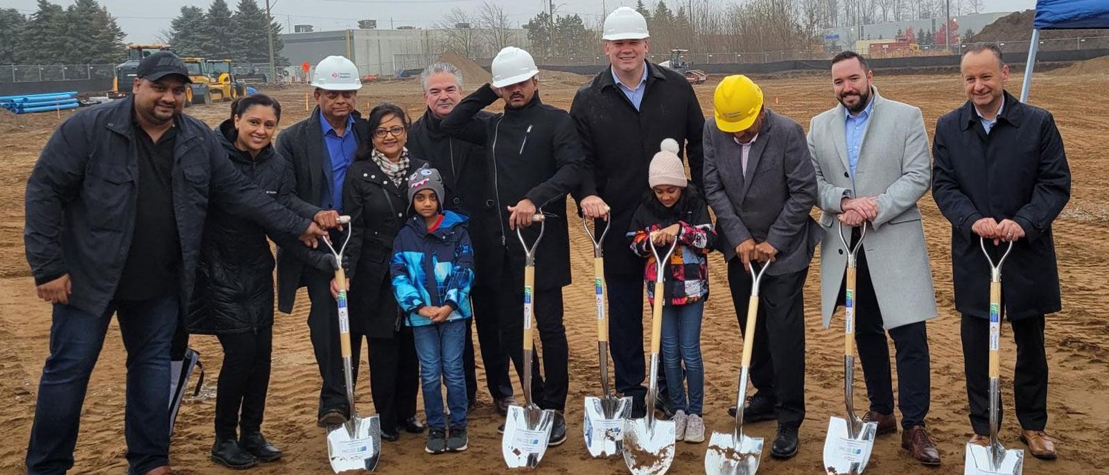 Row of people posing for a ceremonial groundbreaking. They are wearing white and yellow construction helmets and are holding silver spade shovels.