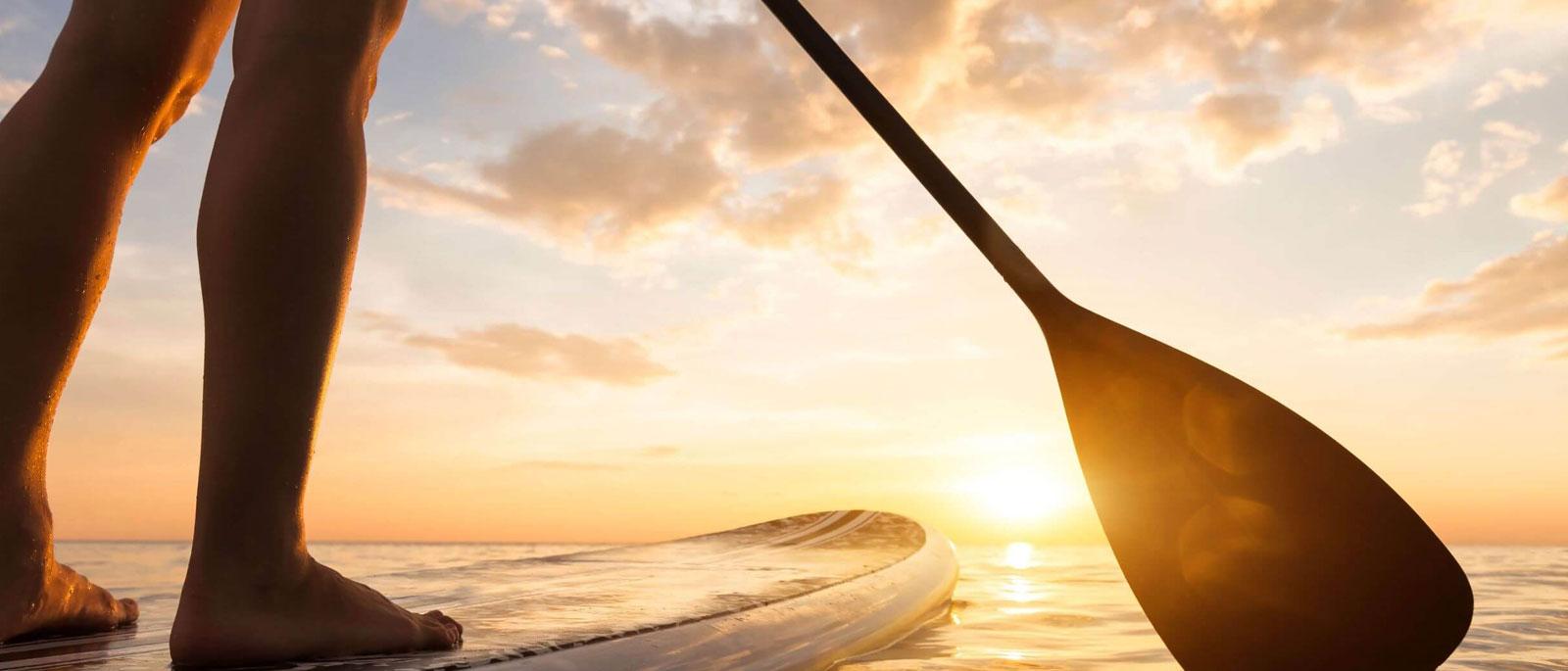 Paddleboarder at sunset with paddle, calm water and glowing sky.