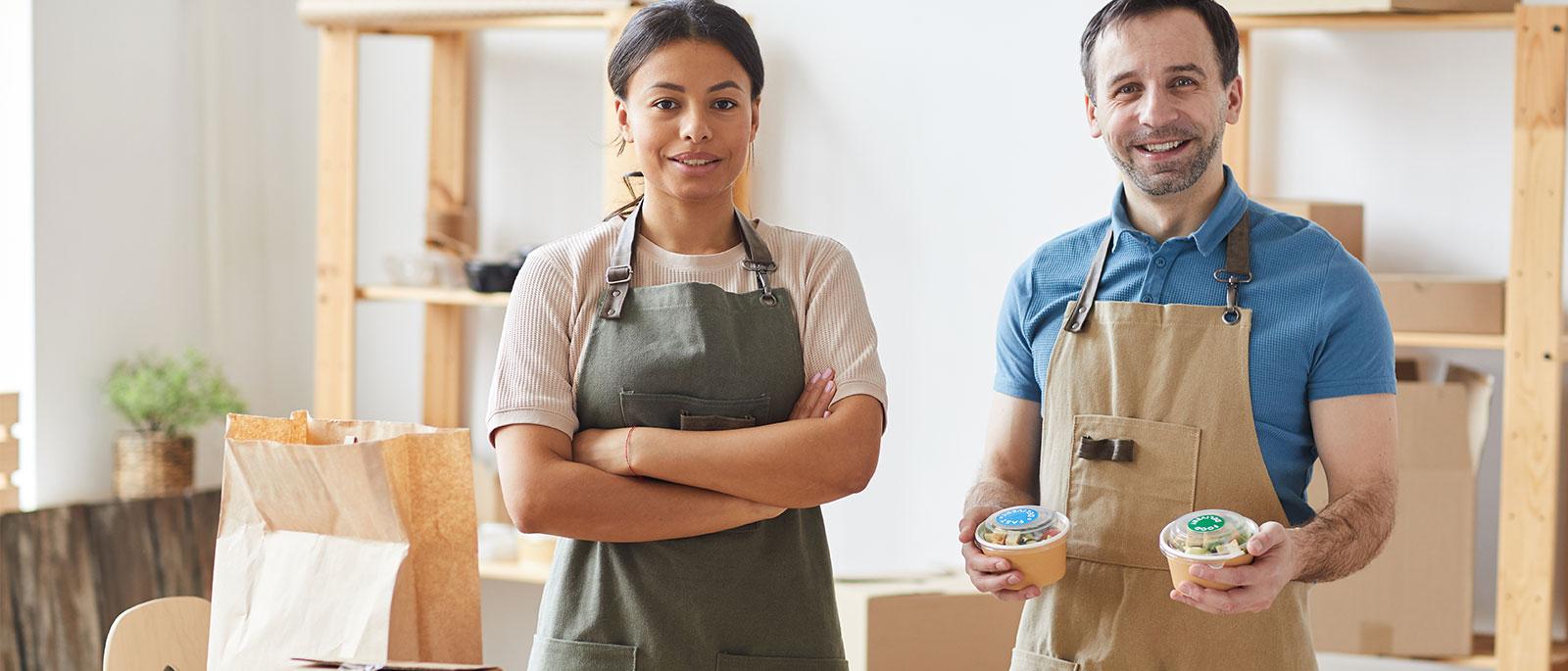 Two people in aprons standing in a kitchen, holding packaged food.