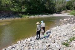 Two people fishing by a rocky riverbank surrounded by trees.
