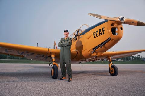 Pilot in green jumpsuit stands in front of a yellow vintage aircraft on a runway.