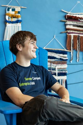 Young person smiling, seated on a blue chair, with woven wall art behind.