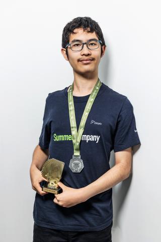 Young person wearing a medal, holding a trophy, against a plain background.