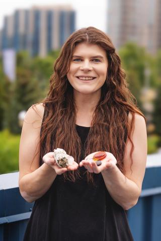 Woman smiling, holding objects in hands, urban greenery in background.