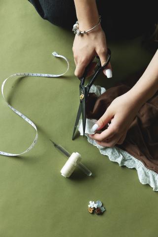 Hands cutting lace with scissors on green fabric, measuring tape and pins nearby.