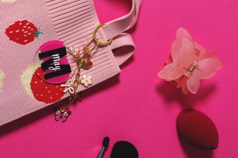 Strawberry-patterned bag, pink butterfly clip, red sponge, and makeup brush on pink background.