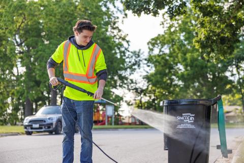 Man in reflective vest power-washes trash bin on a sunny street.