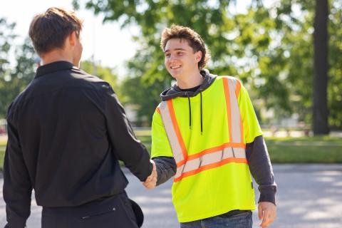 A young man in a safety vest shakes hands with another person outdoors.