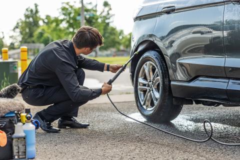 Man washing car tire with hose, bottles nearby.