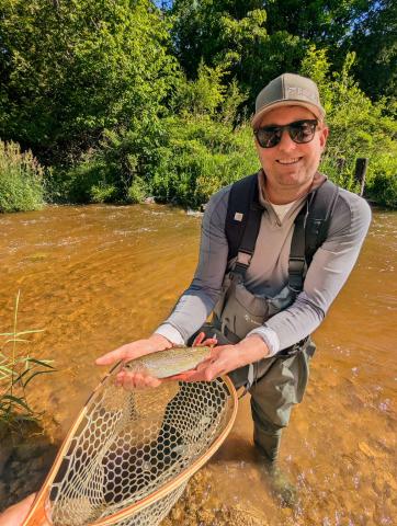 Man in waders holding a fish over a net in a sunny, shallow stream.