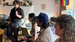 Person standing and gesturing during a discussion in a cozy living room setting with three seated listeners.