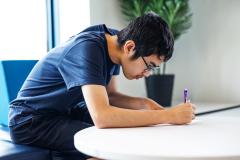 Teen writing at a table, focused, with a plant in the background.