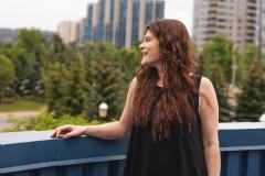 Woman with long hair standing on a balcony, cityscape in background.
