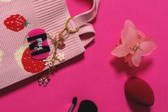 Strawberry-patterned bag, pink butterfly clip, red sponge, and makeup brush on pink background.