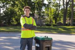 Young man in a safety vest stands next to a recycling bin on a sunny street.