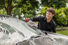 Smiling person washing car windshield with sudsy sponge outdoors.