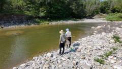 Two people fishing by a rocky riverbank surrounded by trees.