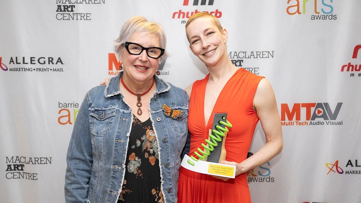 Two people standing in front of a backdrop with logos for Barrie Arts Awards, MacLaren Art Centre, Rhubarb Media, Allegra Barrie and MTAV. One of the people is holding an award.