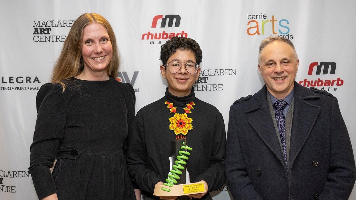 Three people standing in front of a backdrop with logos for Barrie Arts Awards, MacLaren Art Centre, Rhubarb Media, Allegra Barrie and MTAV. One of the people is holding an award. 
