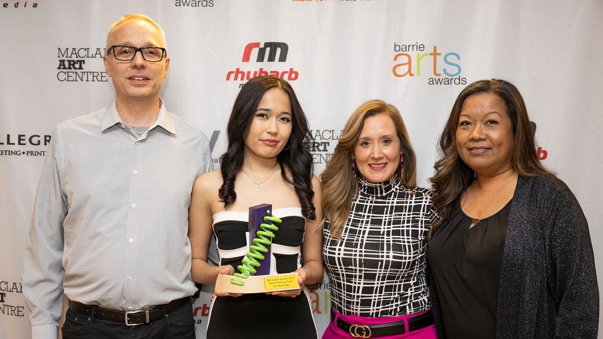 Four people standing in front of a backdrop with logos for Barrie Arts Awards, MacLaren Art Centre, Rhubarb Media, Allegra Barrie and MTAV. One of the people is holding an award. 